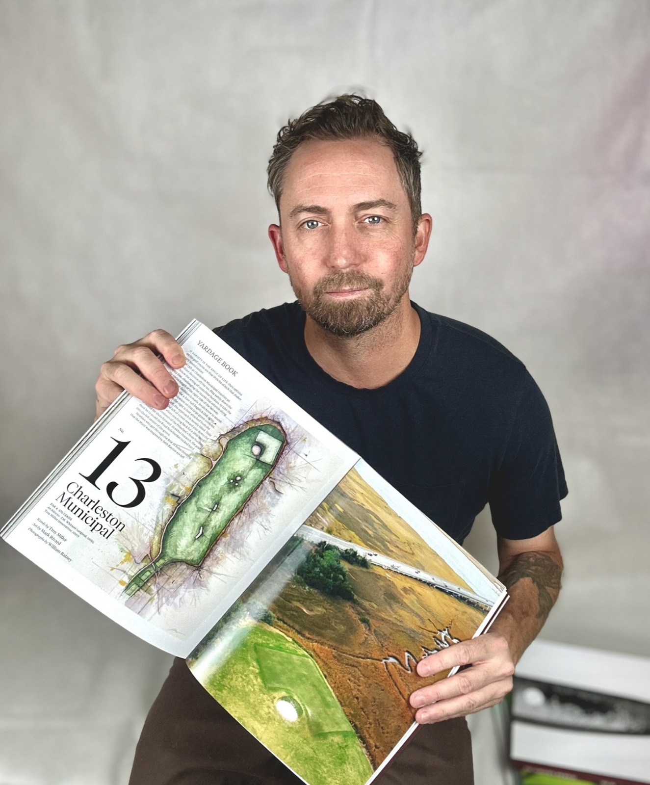 Artist Mark Rivard holding a copy of The Golfer's Journal open to the yardage-book spread featuring his painting of Charleston Municipal hole No. 13