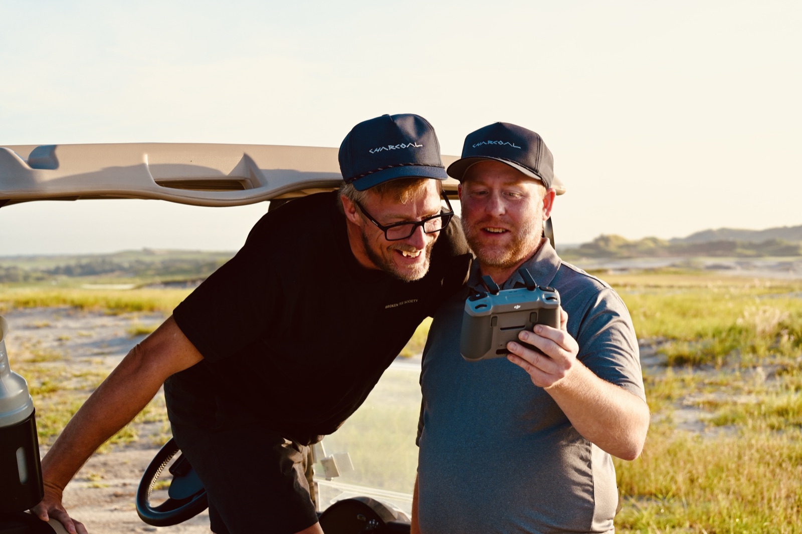 Charcoal Golf founders Patrick Jacobson and Russ Olson in Charcoal caps, reviewing drone footage beside a golf cart